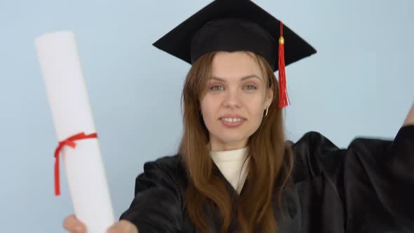 Young Caucasian Female Student in a Black Gown and Master's Hat Holds in Her Hands in an Upright