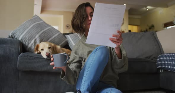 Caucasian woman reading documents, working from home with her pet dog on sofa next to her