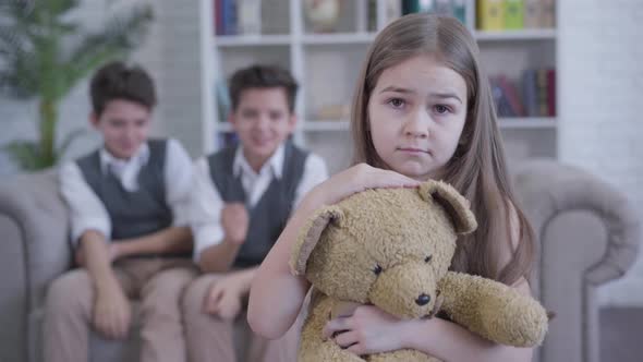 Sad Pretty Caucasian Girl with Teddy Bear Looking at Camera As Twin Brothers Pointing on Her 