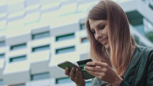 Smiling Young Female Customer Holding Credit Card and Smartphone, on Modern Home