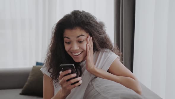 Black Young Woman Looks Into a Smartphone and is Pleasantly Surprised and Happy