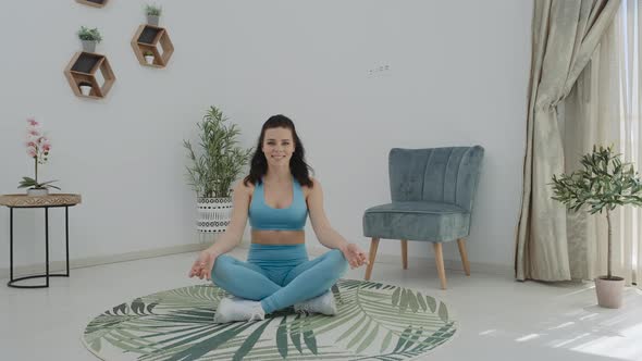 Relaxed brunette woman in blue sportswear meditating in lotus position sitting on carpet breathing