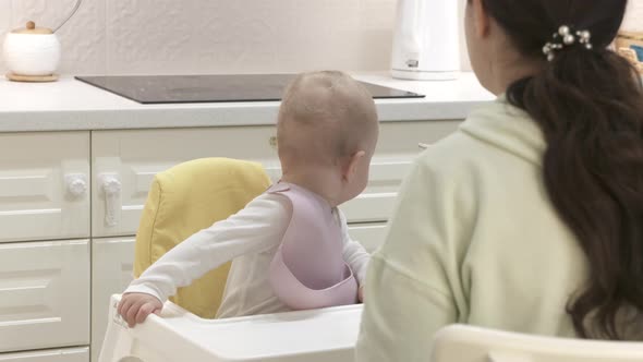 Mother Feeding Oneyearold Baby Boy with a Spoon and a Bowl in the Kitchen at Home