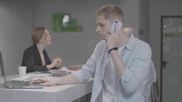 Young Stressed Man Yelling on the Phone and Gesturing with Colleagues Working at the Background