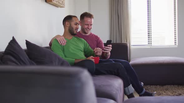 Multi ethnic gay male couple sitting on couch looking at smartphone together