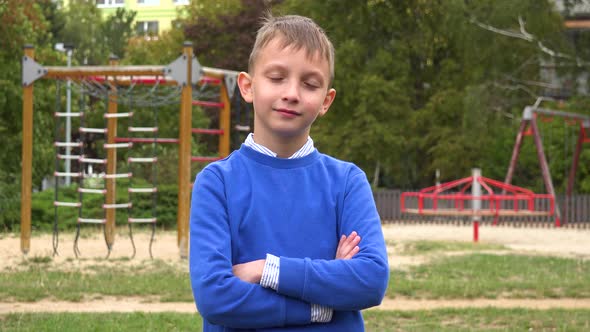 A Young Boy Folds His Arms Across His Chest and Smiles at the Camera in Front of a Playground