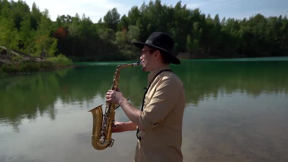 Male Saxophonist Is Playing Melody on Shore of Picturesque Forest Lake at Summer Day