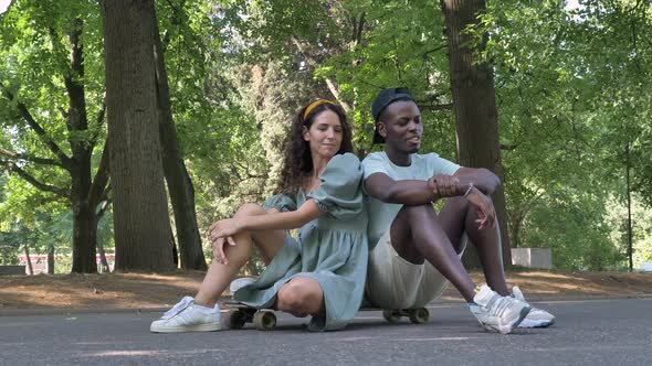 Black Man Talking to His European Girlfriend in a Dress While Sitting on a Skateboard in a Park in