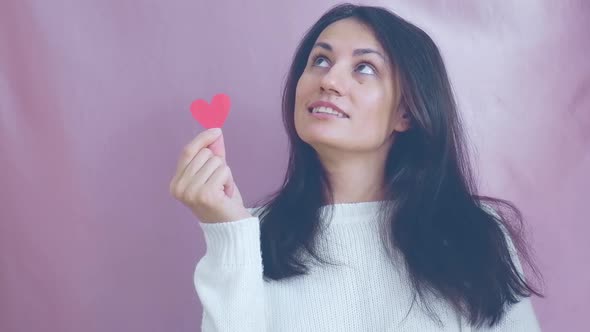 Smiling Funny Young Brunette in a White Sweater Smiles Blinks and Waving Red Hearts on a Pink