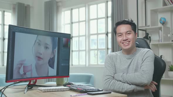 Asian Cameraman Crossing His Arms And Smiling To Camera While Editing Photo By Desktop Computer