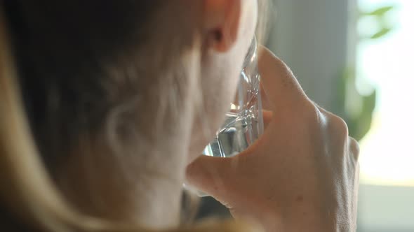 Young Woman with Pigtail Drinks Water From Transparent Glass Against Background of Window During Day