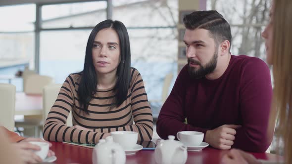 Beautiful Caucasian Couple Sitting in Cafe and Chatting with Blurred Friends. Brunette Man and Woman