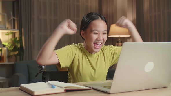 Asian Kid Girl Sitting In A Wheelchair While Using Laptop Computer And Celebrating At Home