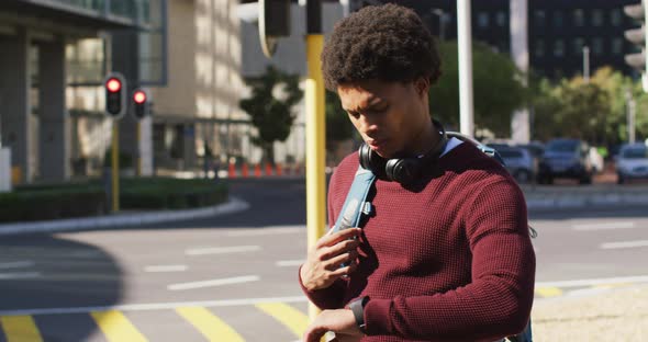 African american man in city checking smartwatch, wearing headphones and backpack waiting in street