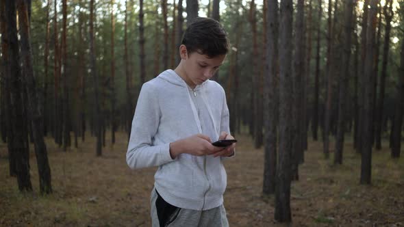 Young Boy Texting on Smartphone Standing on Pine Woods