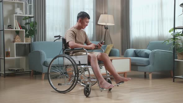 Young Asian Boy Sitting In A Wheelchair While Smiling And Using Smart Phone At Home alt