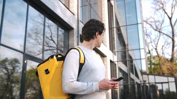 Portrait of Caucasian Delivery Man Standing with Yellow Backpack Using Navigation App on Phone in