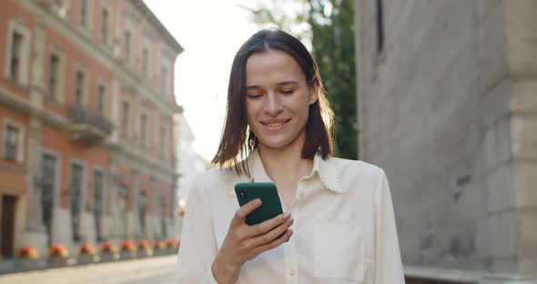 Close Up View of Happy Young Woman Using Mobilephone While Walking Outdoors. Beautiful Girl Smiling