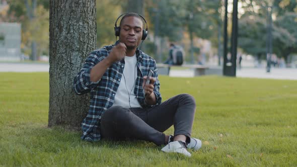 Afro American Guy Sits in Park on Grass in Summer Day Listening Music on Headphones Sings Song