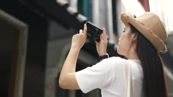 A beautiful young Asian traveler wears a vintage hat using a smartphone taking a photo .