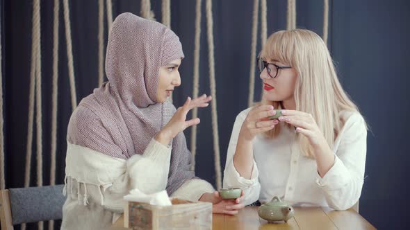 Two Muslim and Caucasian Women Are Chatting Sitting in Cafe and Drinking Tea