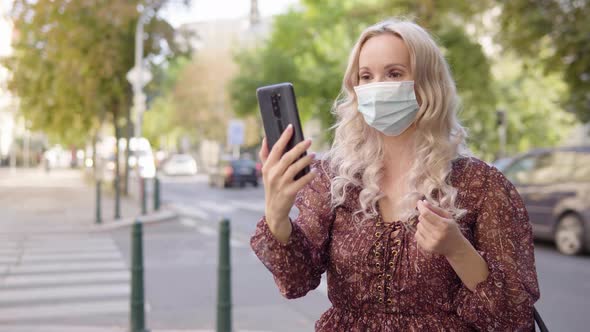 A Middleaged Caucasian Woman in a Face Mask Talks on a Video Call on a Smartphone in an Urban Area