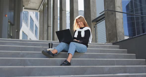 Girl in Casual Clothes Sitting on Steps of Office Building and Uses Her computer