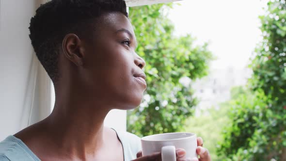 African american woman holding coffee cup looking out of window at home
