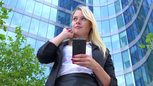 A Young Beautiful Businesswoman Works on a Smartphone - Closeup From Below - an Office Building