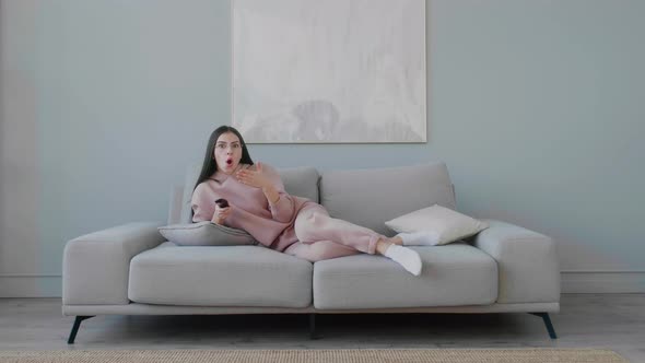 A young woman with glasses is lying on the sofa, yawning, smiling and changing TV channels.