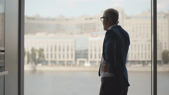 Senior Entrepreneur Standing Near Panoramic Window of Office Looking on Cityscape Talking on Phone