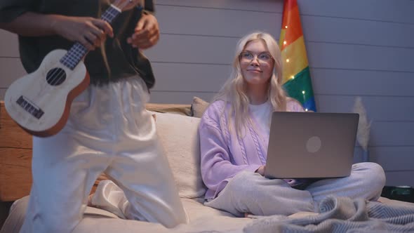 Girl Comes to Her Girlfriend Sittingin Bed Working on a Laptop to Play Ukulele