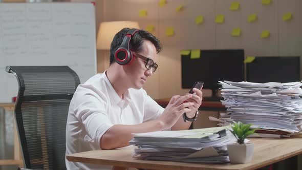 Asian Man With Headphones Listening To Music On Smartphone After Working With Documents