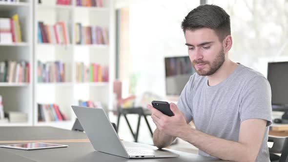 Young Man Using Smartphone and Laptop in Office