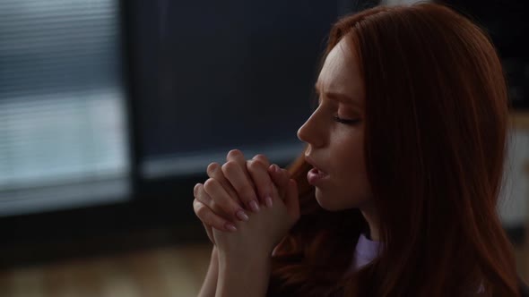 Closeup of Depressed Young Woman Praying with Eyes Closed Holding Folded Hands in Front of Face at