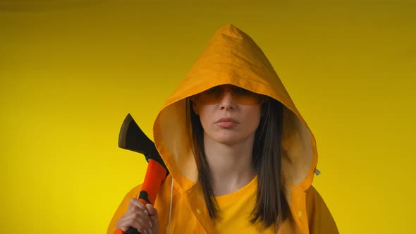 Young Woman in a Yellow Raincoat Holds an Axe in Her Hand in a Yellow Studio