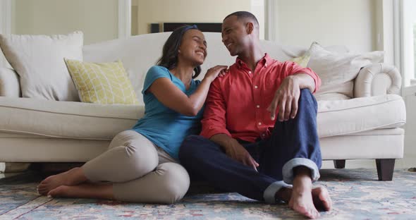 Happy biracial couple sitting on floor in living room, embracing and looking at camera