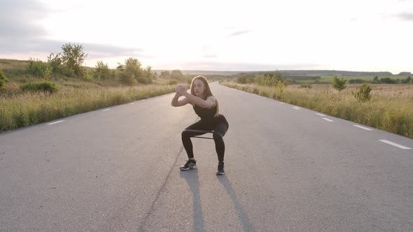 The Girl Training Squats with Fitness Elastic Band on Road at Nature with Sunset