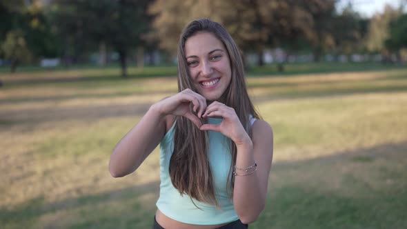 a Cute White Girl Makes a Heart with Her Fingers in a Park in the City