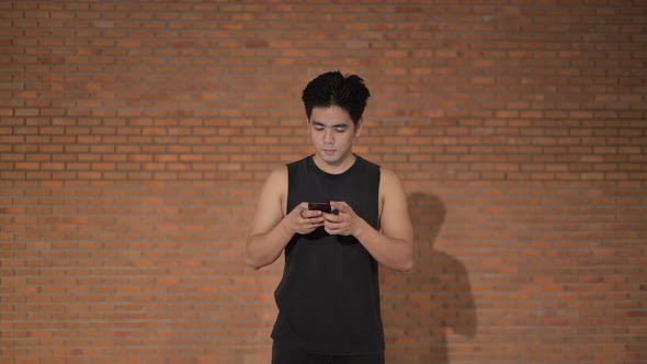 Young Happy Handsome Asian Man Thinking While Using Phone Against Brick Wall