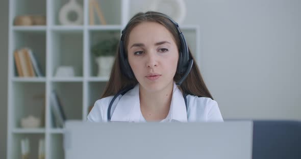 Female Medical Assistant Wears White Coat, Headset Video Calling Distant Patient on Laptop. Doctor