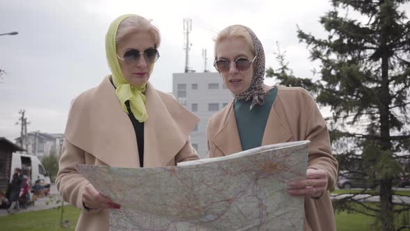 Two Mature Women in Headscarves and Sunglasses Holding the Map and Talking. Senior Ladies Travelling