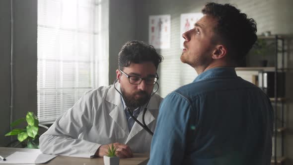 Focused Young Male Cardiologist Listening Heartbeat of Young Male Patient at Check Up Meeting in