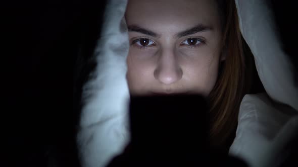 A Young Woman Lies Under a Blanket with a Phone in Her Hands. A Woman in the Dark Uses a Pad. Modern