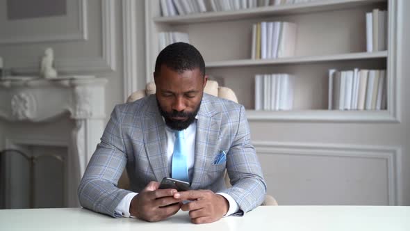 Tired Rich Man Is Checking News in Internet By Mobile Phone Sitting at Table in His Home, Leaning on