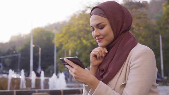 A Young Beautiful Muslim Woman Looks at a Smartphone with a Smile in a Street in an Urban Area