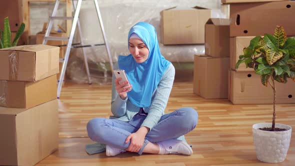 Portrait Young Muslim Woman in a Hijab Uses Smartphone Sitting on the Floor Next To the Boxes in a