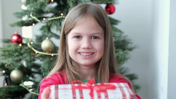 Cute Little Girl Smiling Giving Christmas Gift Box at Home Near Christmas Tree