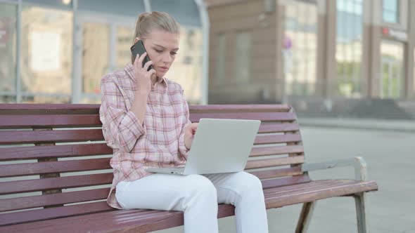 Young Woman Talking on Phone and Using Laptop While Sitting Outdoor on Bench