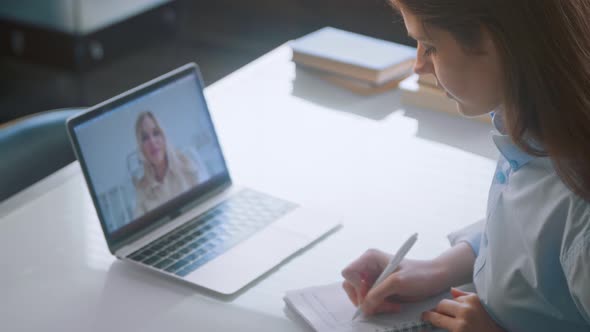 Young lady doctor makes notes in paper notebook listening to patient at online consultation